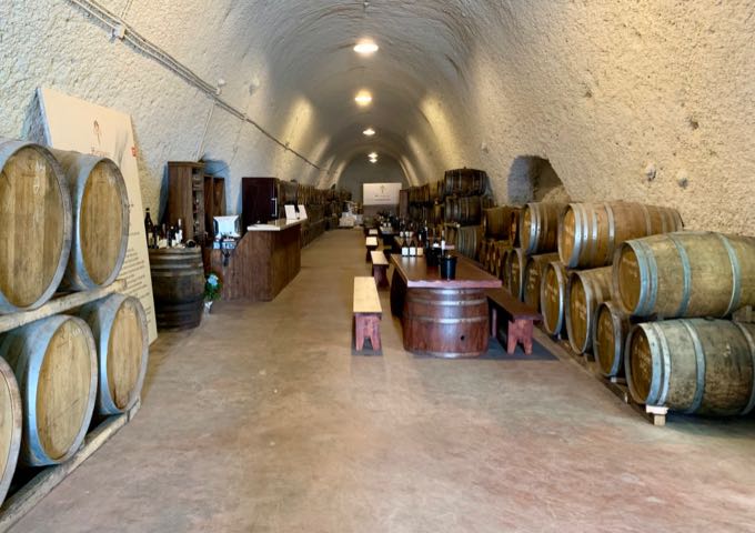 Long narrow cave room lined with wine barrels, with a table set up for tasting