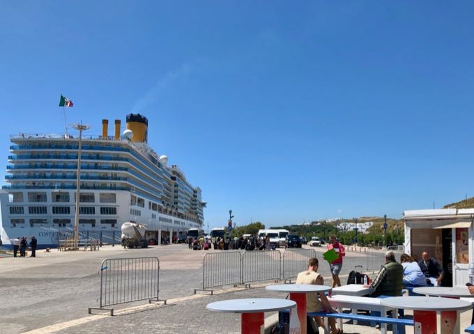 A cruise ship is docked at Mykonos New Port, while travelers wait at picnic tables in front of the terminal.