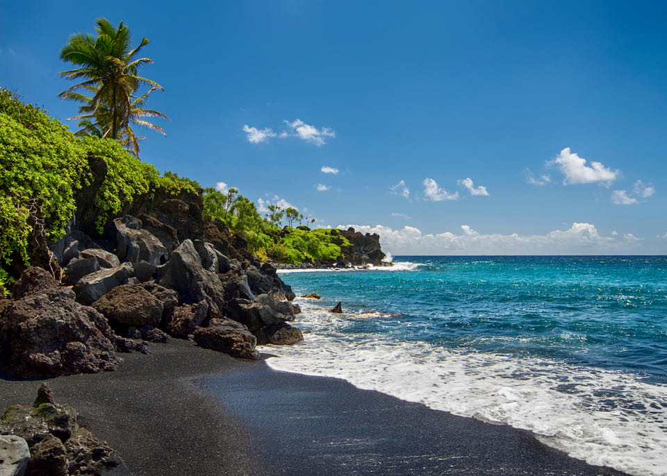 Beach with black sand, turqoise water, and rocky terrain with lush vegetation