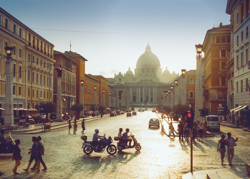 Šiuolaikinė Romos gatvė saulėlydžio metu, pėstieji ir žmonės ant motorolerių, o Šv. Petro bazilika fone.