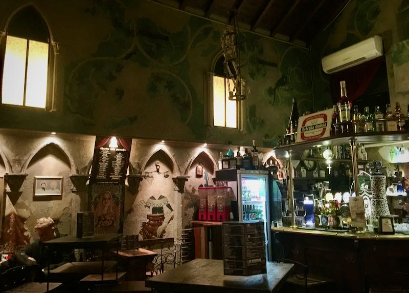 Dimly-lit interior of a bar with arched Gothic windows and frescoes on the wall