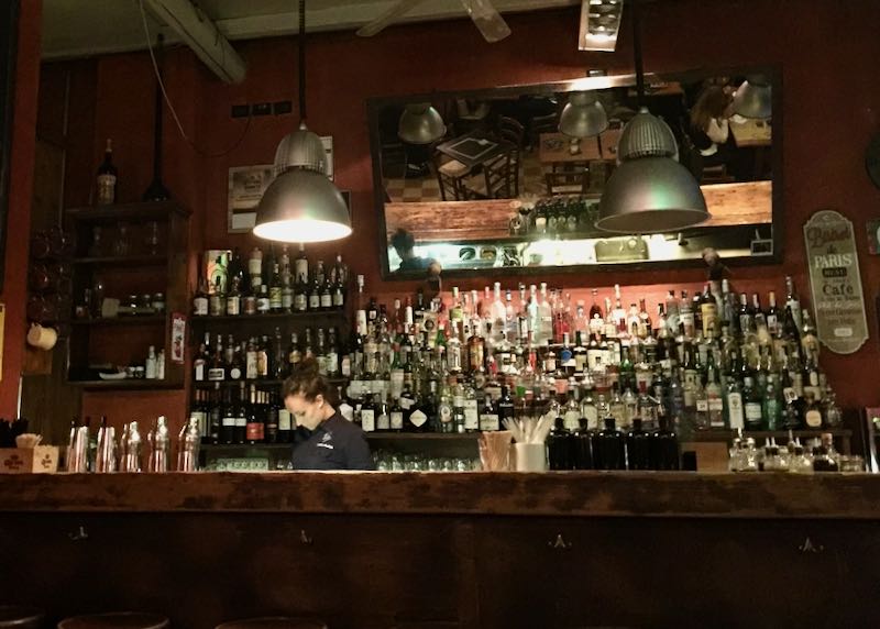 A female bartender stands in front of shelves of bottled spirits
