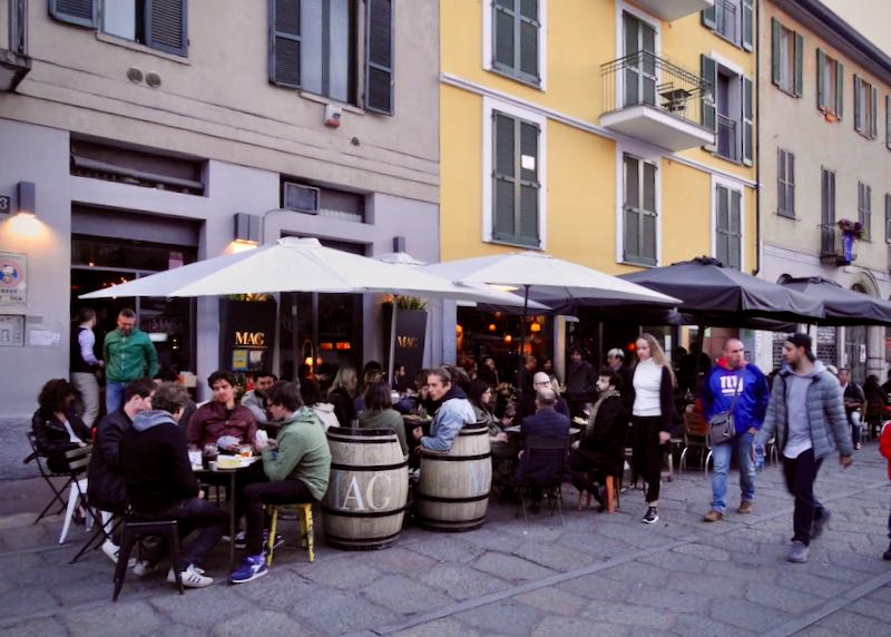 Patrons seated under umbrellas at tables made from old wine barrels on a sidewalk terrace in Milan