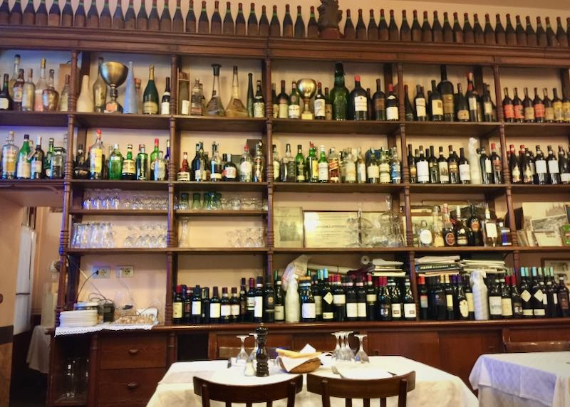 Two wooden chairs and a table are set before shelves of bottles in an Italian restaurant