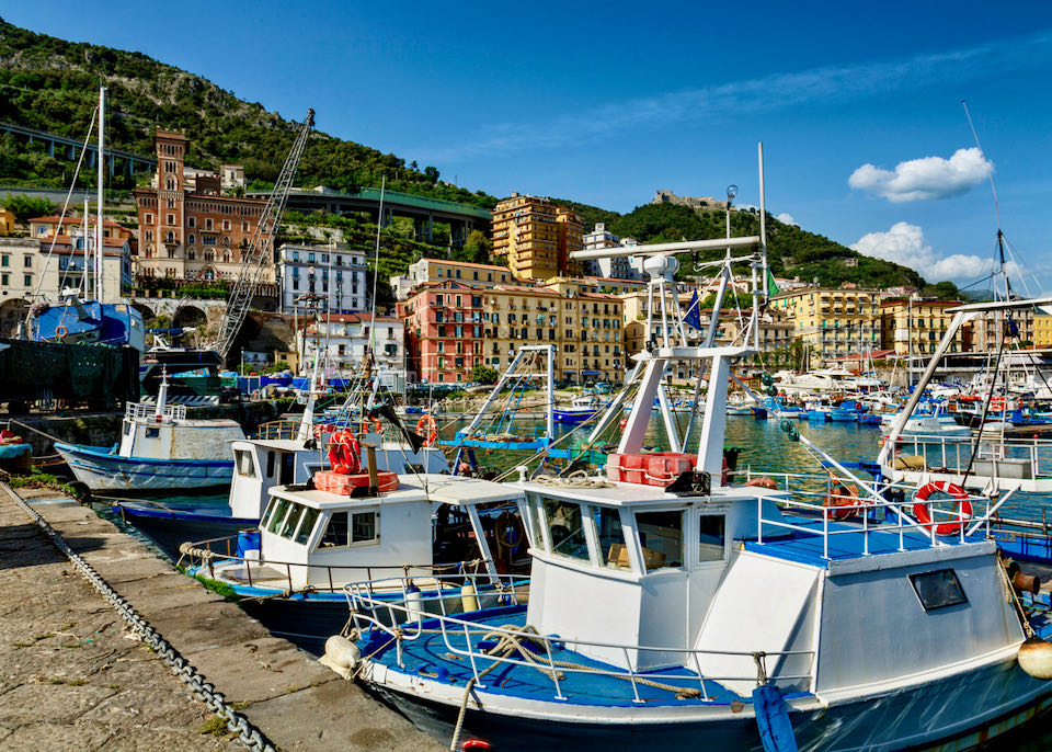 Colorful fishing boats line a harbor, with Venetian-style buildings and tree-covered hills in the background