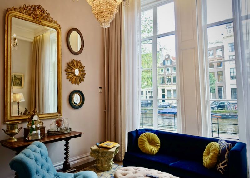A blue velvet sofa sits in front of a curtained window that overlooks an Amsterdam canal