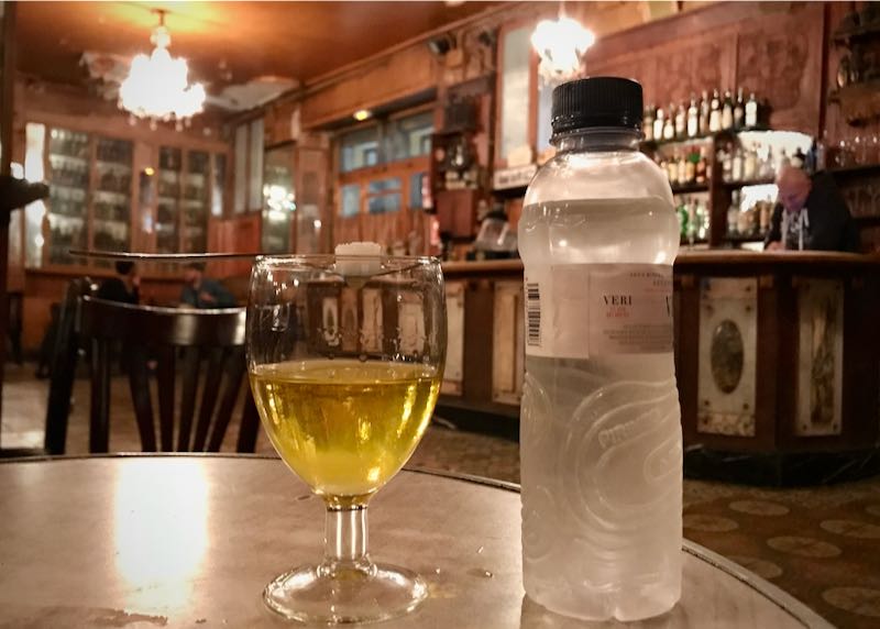 A glass of white wine and a bottle of water sit on a bar table, with a bartender in the background