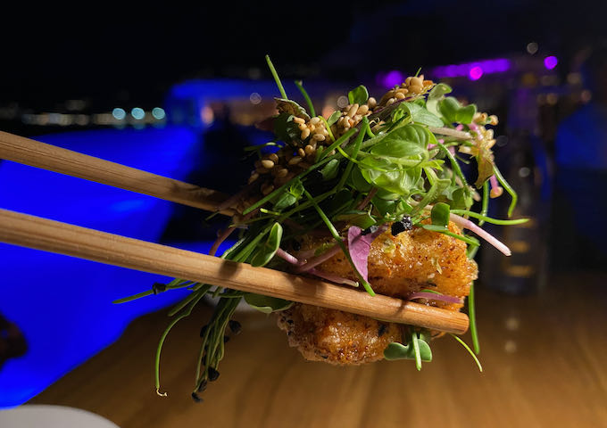 A bite of tempura-fried food is held aloft with chopsticks with a under-lit swimming pool in the background