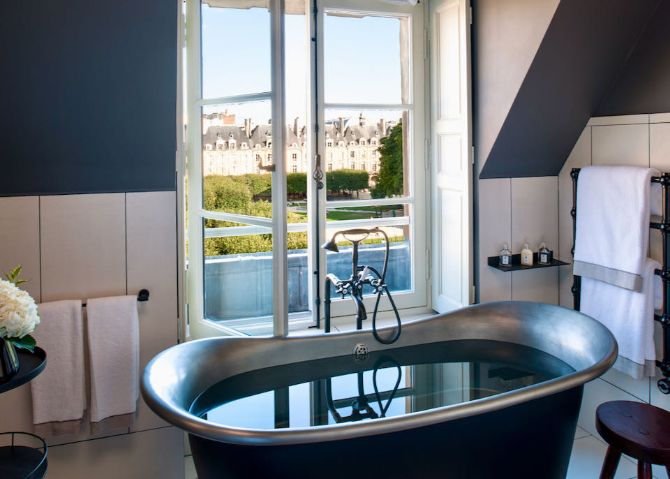 Clawfoot bathtub in front of an open window with views of a leafy Parisian square