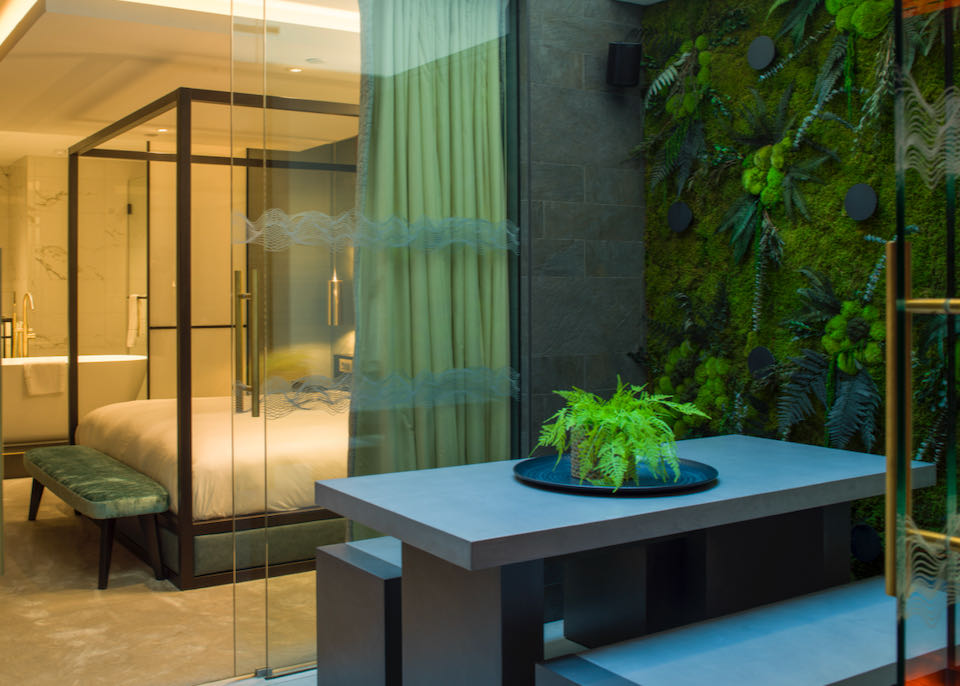 View from a hotel balcony with green "living wall" into a hotel room with a modern four-poster bed and pedestal bath