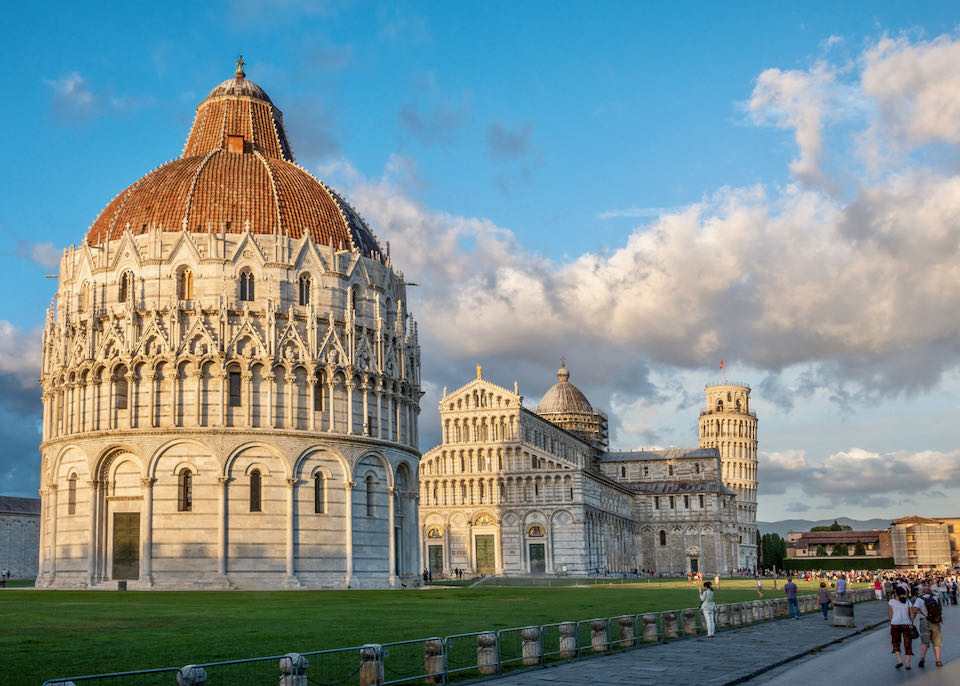 Photo of San Giovanni Baptistery, Pisa Cathedral, and Leaning Tower of Pisa at sunrise, with tourists taking photos.