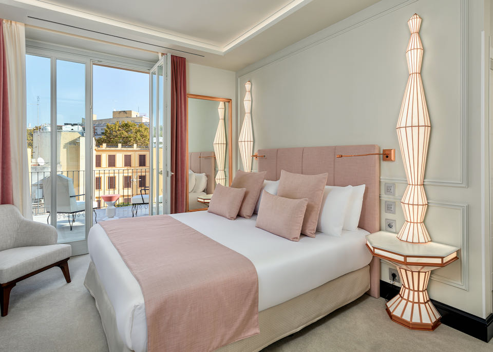 Hotel bed with padded headboard and linens in cream and blush tones, next to an open balcony door looking over Rome