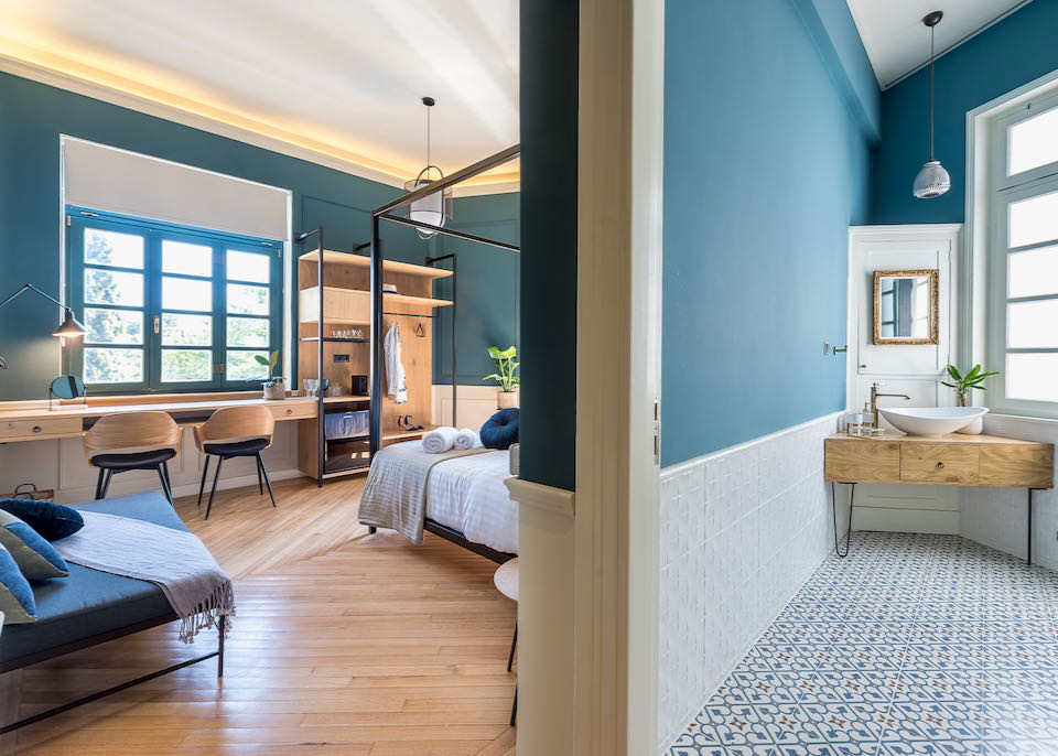 Image taken from a doorway, with a hotel bedroom on one side and bathroom on the other. Both have high ceilings, modern decor, and stylish teal paint.