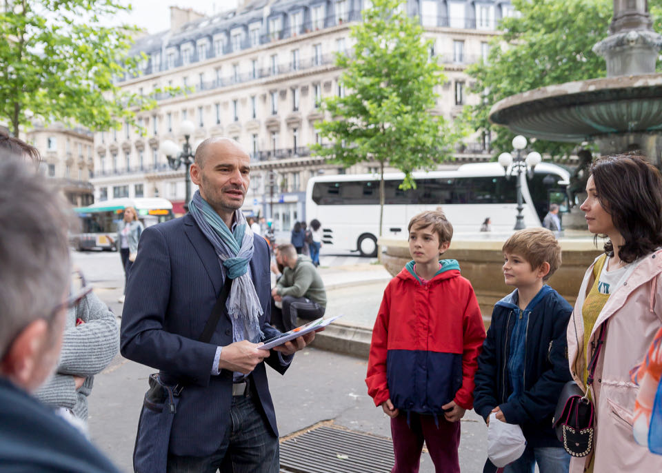 A dapper-dressed man speaks to a small crowd of tourists in a Paris neighborhood.