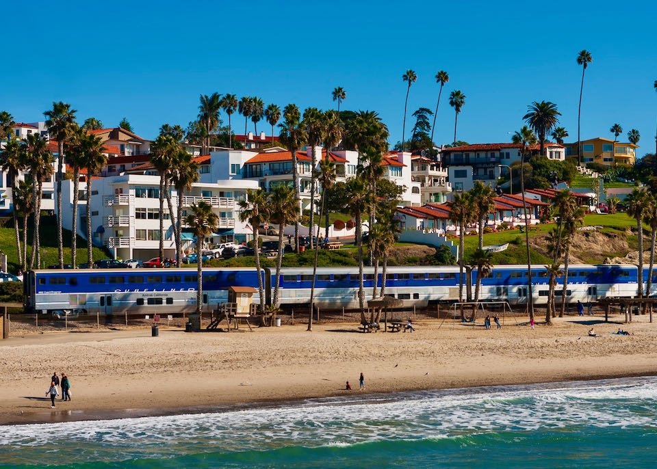 A train travels along a sandy beach lined with palm trees and red-roofed, mission-style buildings.