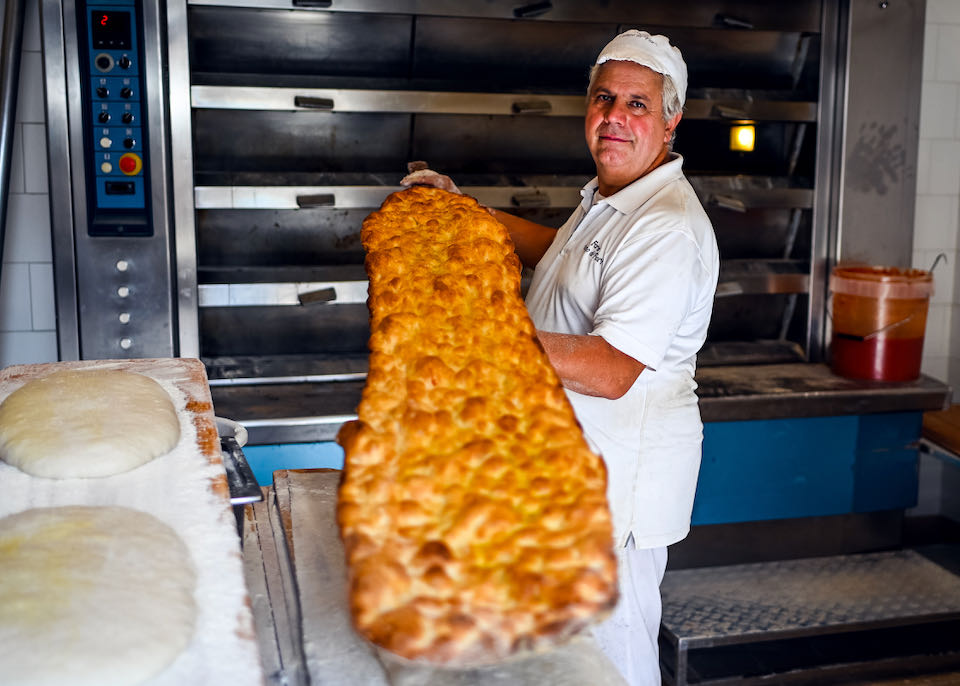 A man in a baker's uniform pulls a huge, onlong pizza from a wall oven and shows it to the camera.