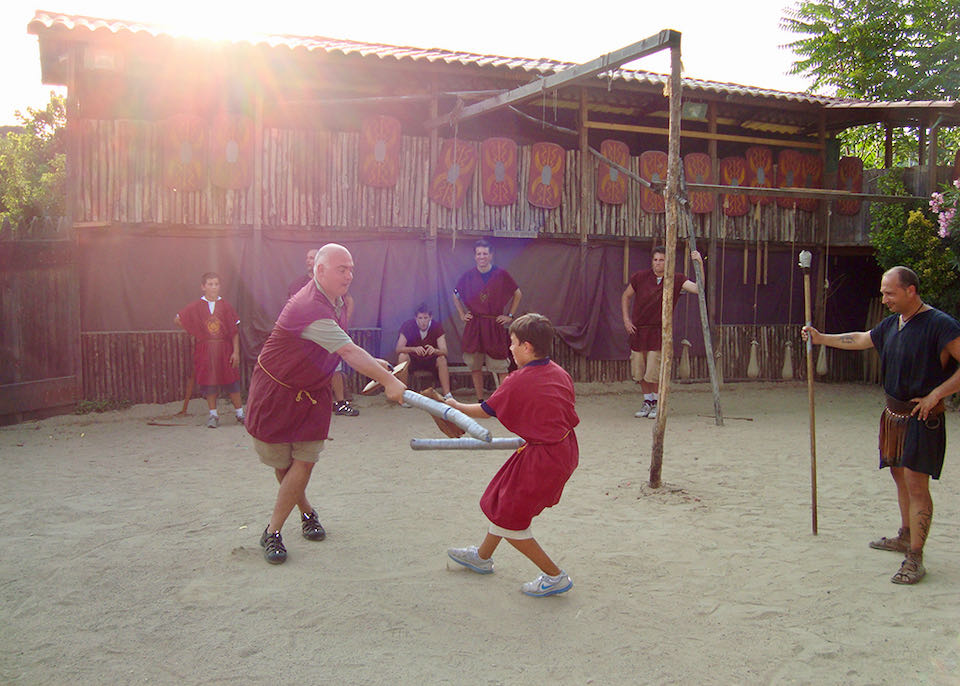 A father and son dressed in ancient Roman costume play-fight in a small dirt arena as others look on