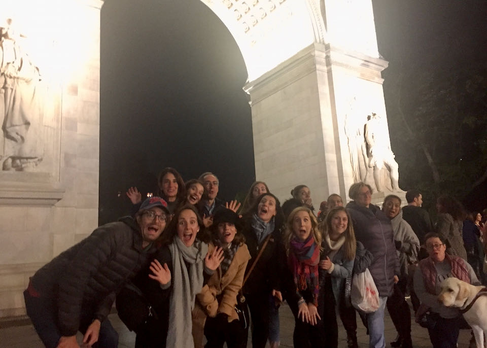 A crowd of people pretend to act scared for the camera, while standing in front of Washington Square Park arch at night