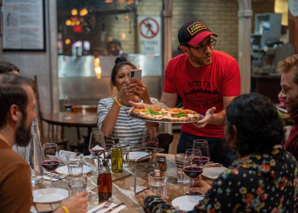 In a pizzaria man serves a large pizza to a table of happy people; one woman is taking a photo with her phone.
