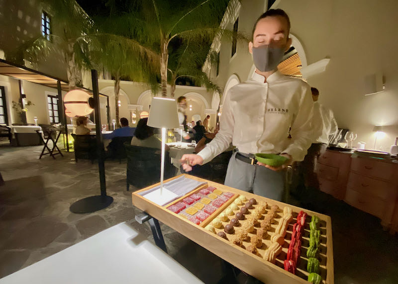 A waiter stands behind a serving tray of desserts inside an arched atrium dining area with palm trees