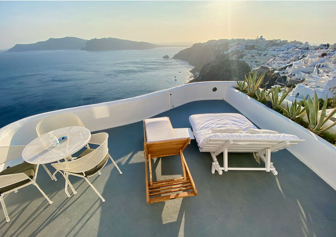 View of the Santorini caldera from a hotel balcony with lounge chairs and a cafe table