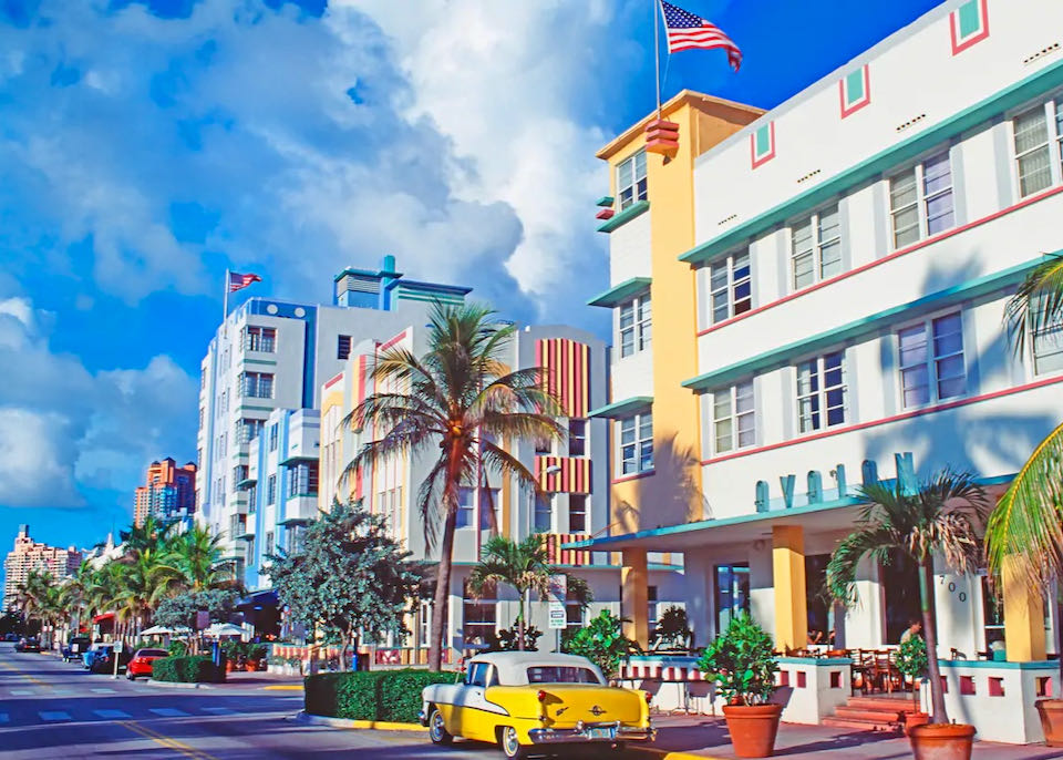 A vintage banana-yellow car sits in front of a boulevard lined with palm trees and colorful art deco buildings on a sunny day.