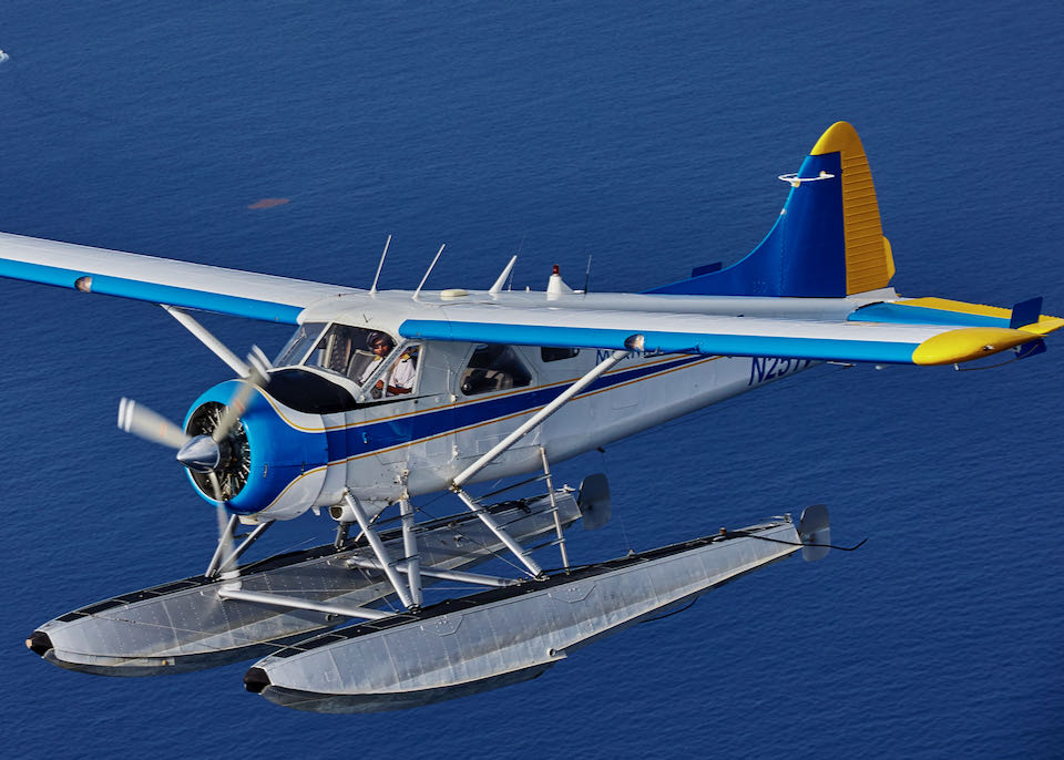 A seaplane with blue and yellow wings flies over open water on a sunny day