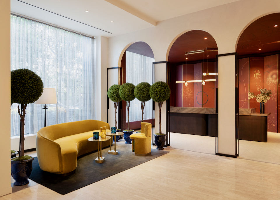 Hotel lobby with plush, mustard-colored sofa and chair, live topiaries, and arched doorways.
