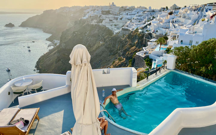 A man in a hat lounges in a cliffside swimming pool with a view over the Santorini Caldera