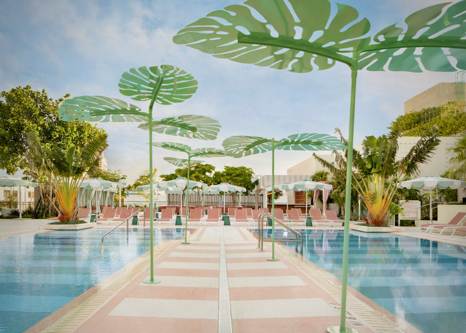 Pool deck with pink tile and cabanas, and large green plastic palm leaves