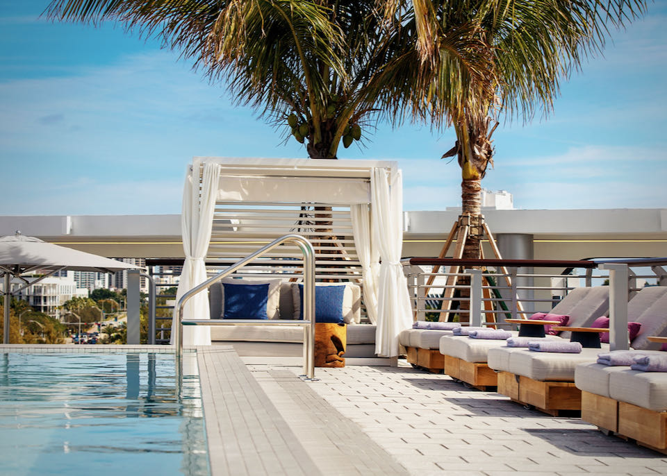 Padded sun beds and a private cabana sit next to a rooftop pool backed by palm trees and a city skyline.