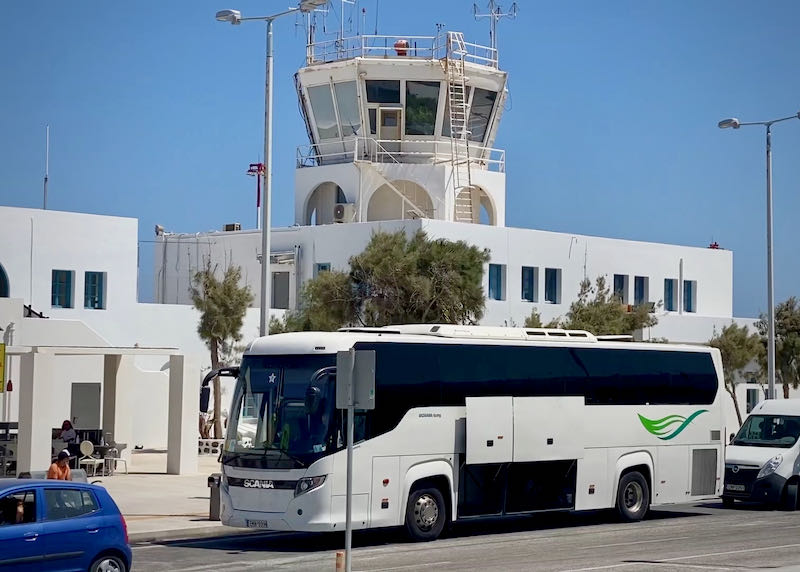 A large, white bus sits outside the Santorini Airport, with the airport control tower in the background. 