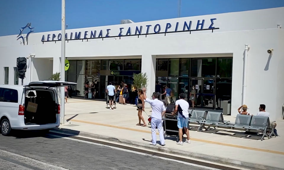 Travelers with luggage enter and exit the white, low-slung building of Santorini Airport