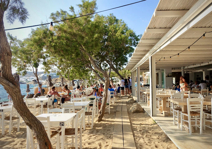 A beachfront restaurant with many tables set in the sand under a beautiful shade tree