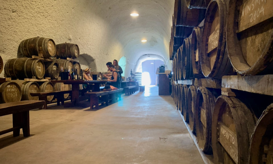 A small group of people drinks wine at a table in a wine cave lined with barrels