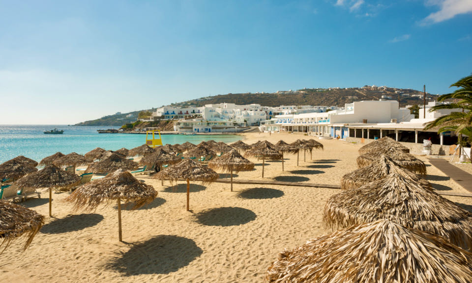 A wooden boardwalk extends over the sand past a row of thatched beach umbrellas and sun beds