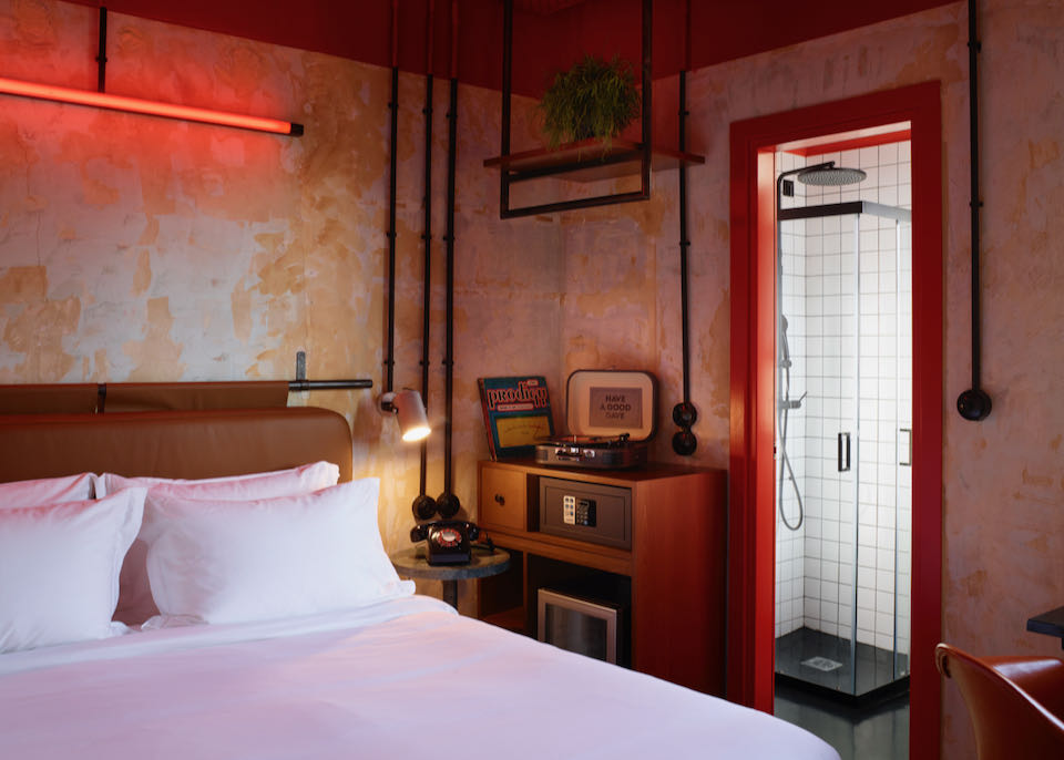 White hotel bed in a room with red neon lighting, a vintage radio and record player, and an ensuite bathroom.