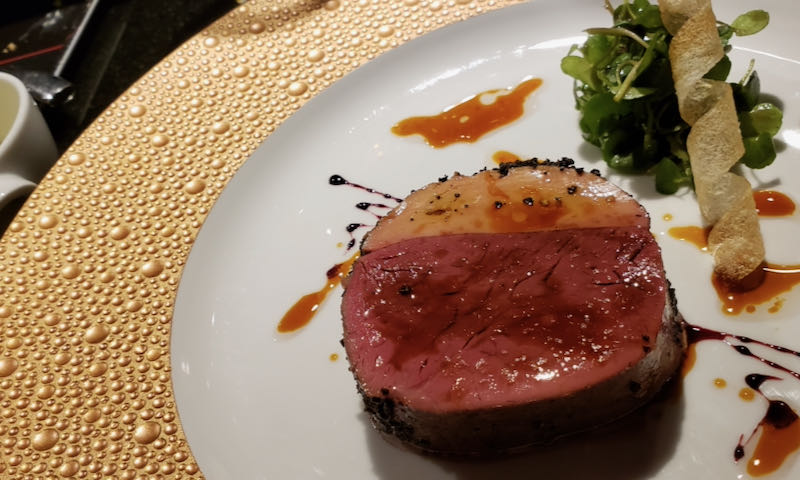A small, seared fillet of meat on a gold-rimmed plate, drizzled with colorful sauces.