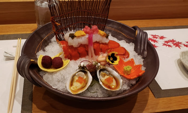 Oysters and sushi rolls sitting atop a bowl of ice, with a decorative flower in the center