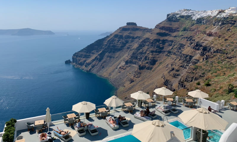 A row of umbrella-shaded sun loungers line a pool at the edge of a caldera-view terrace on a Santorini cliffside.