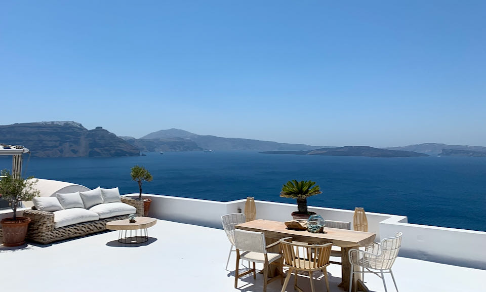 A white furnished terrace overlooking the blue Aegean sea and volcanic islands.