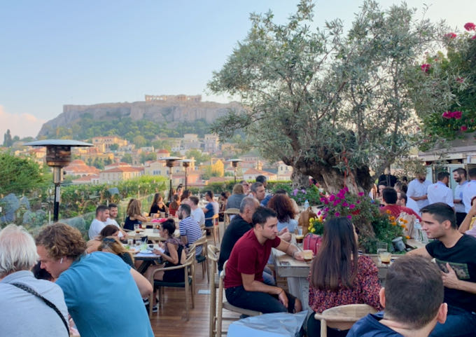 Diners at outdoor tables on a garden terrace with the Acropolis in the background.