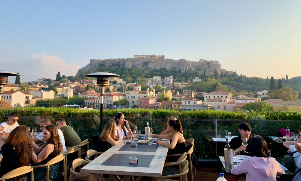 A group of female diners sit at an outdoor table with the Acropolis and Parthenon in the distance.