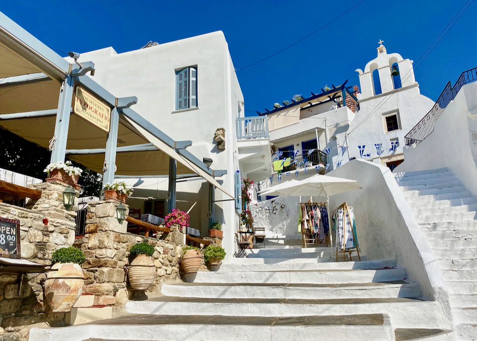 A white stone staircase lined with flower pots leads past a rustic restaurant to a white Greek church.