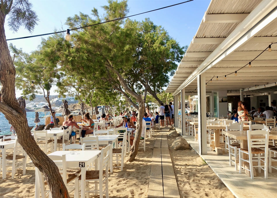 Diners sit at tables in the sand at a beachfront restaurant shaded by trees