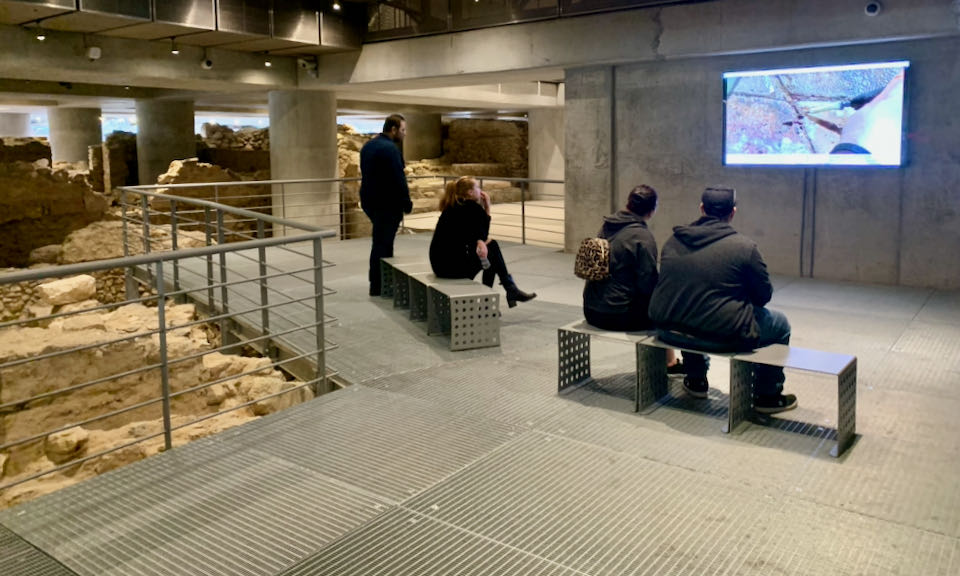People sit on a bench in an archaological site and watch a video