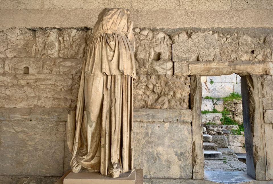 Marble statue of a robed woman, missing arms and head, next to a stone wall and doorway