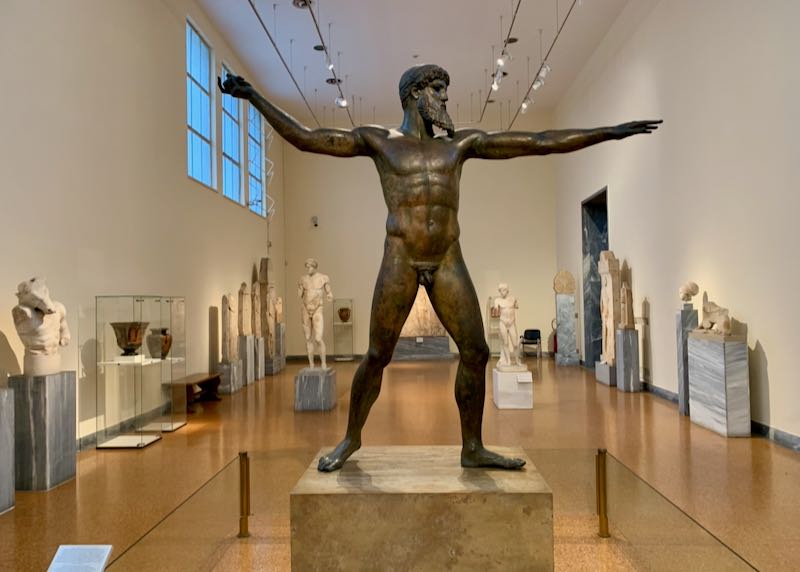 Bronze statue of a man holding a spear in a large museum gallery filled with sculptures