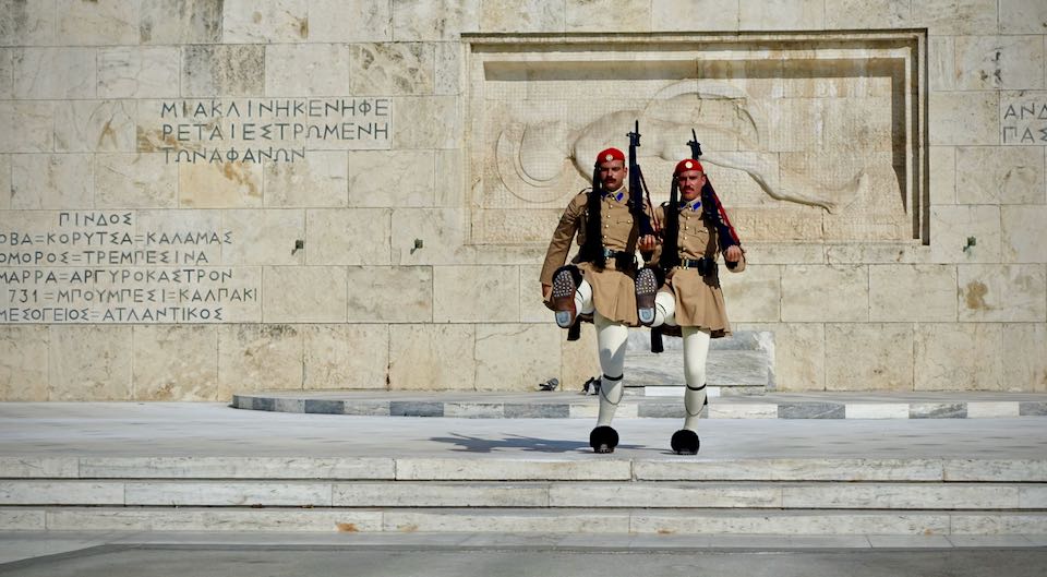 Two men in Traditional Greek military uniforms high-step in tandem.