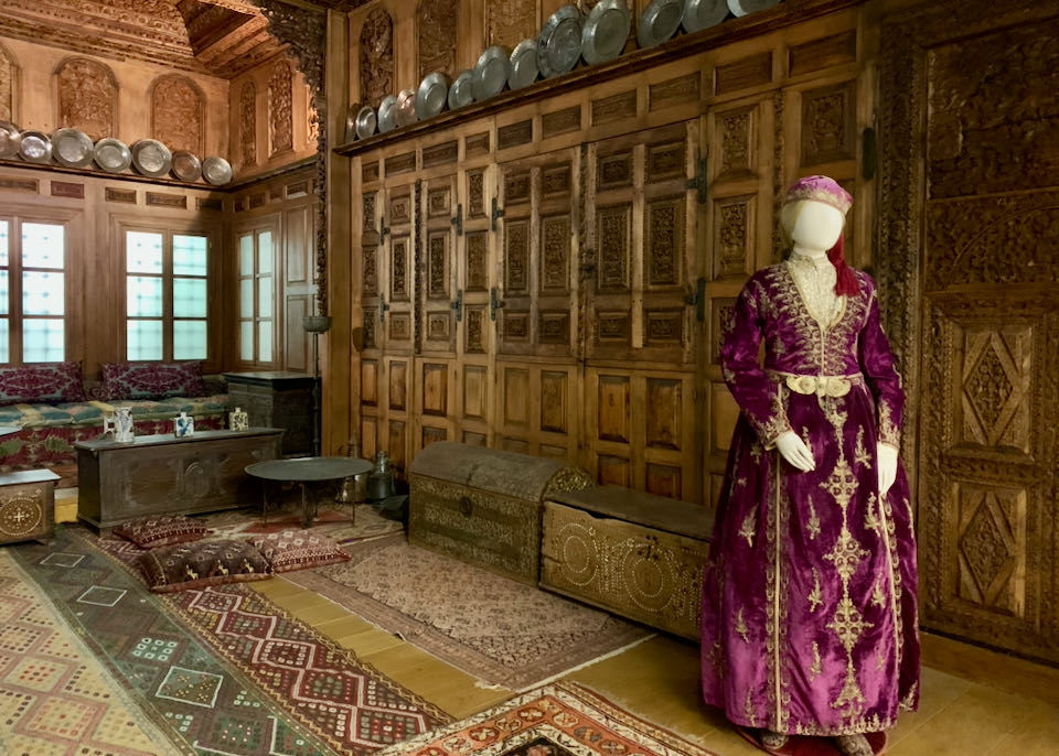A mannequin in elaborate historic costume is displayed in a room of carved wooden panels and colorful textiles